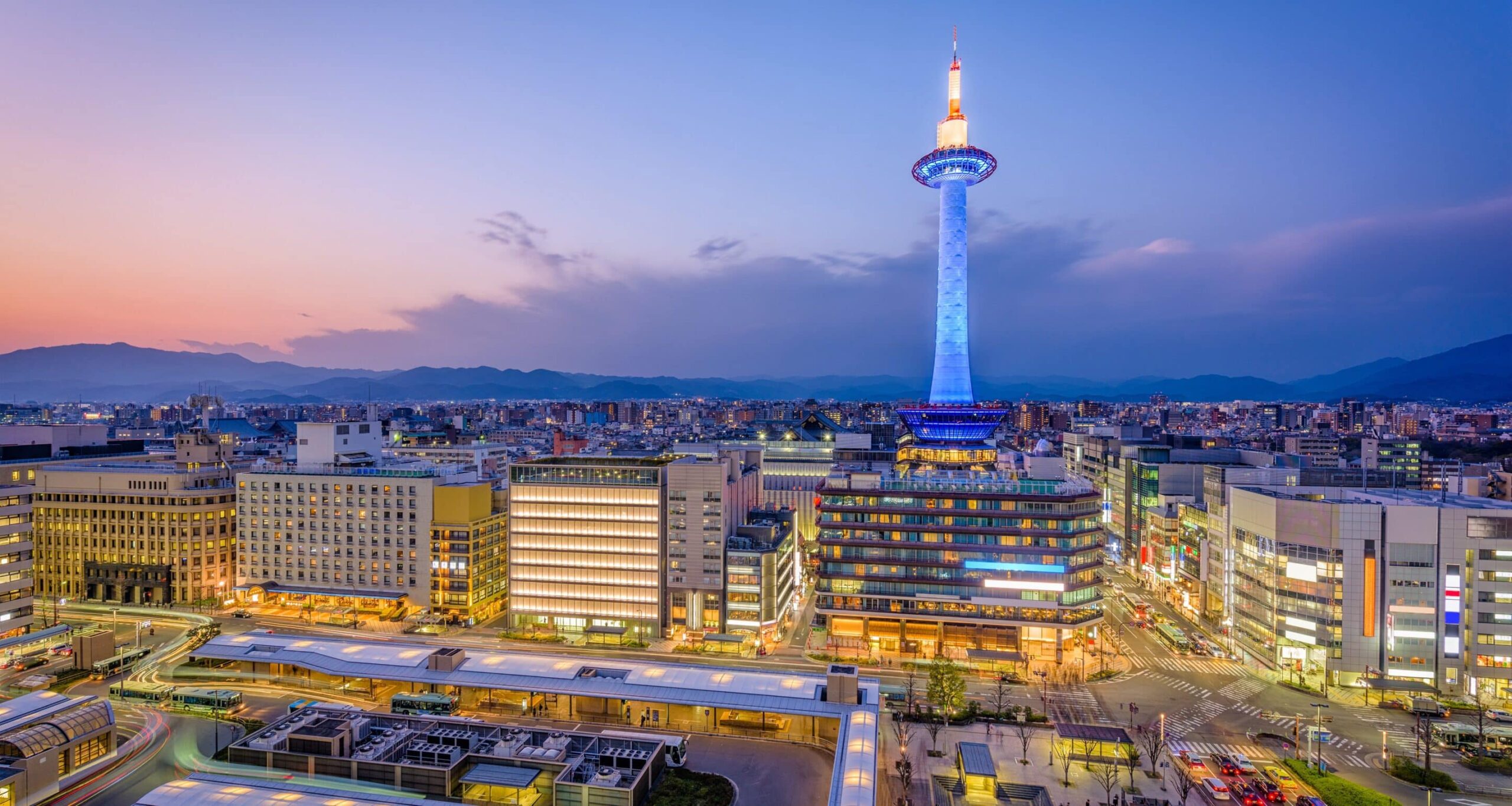 Cityscape at dusk: tall tower with glowing blue top, modern buildings, skys warm and cool gradient, mountains in the background.