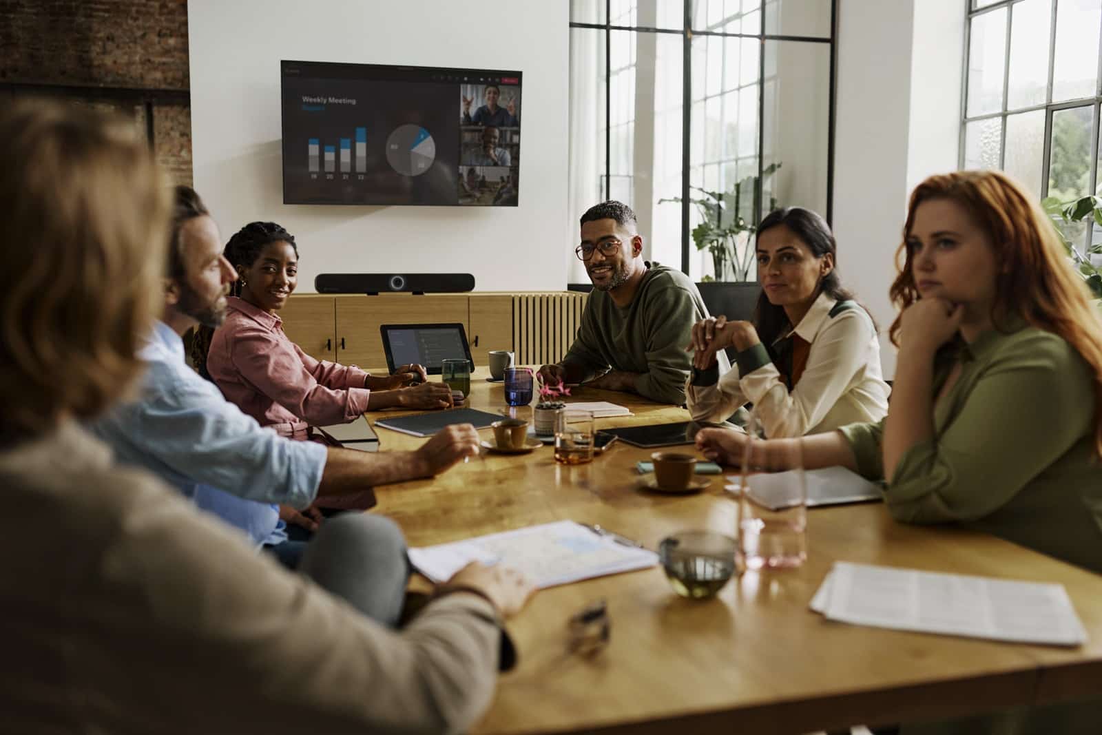 A diverse group sits at a conference table with a presentation screen, papers, laptops, cups, and natural light streaming in.