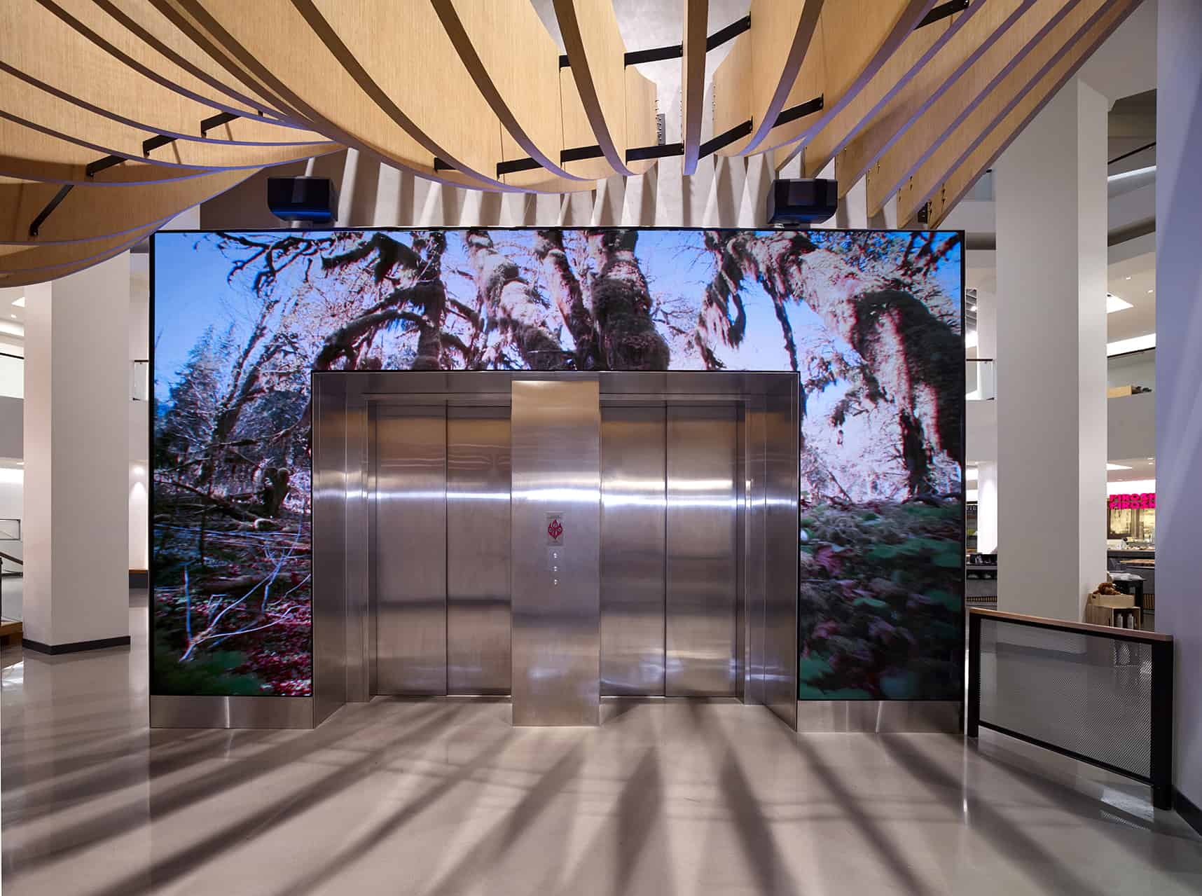 Stainless steel elevator doors with a digital forest display; wavy wooden ceiling casts shadows on the polished floor.