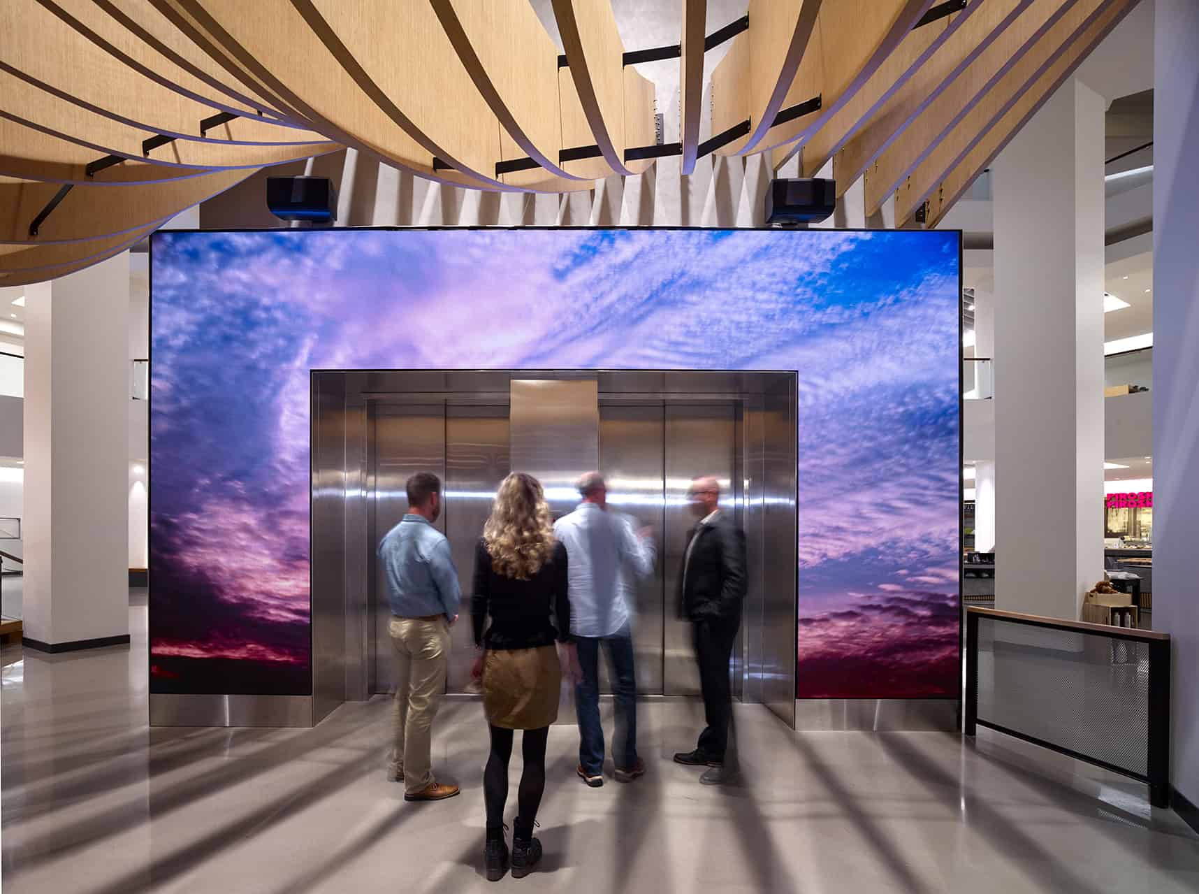 Five people stand by an elevator with a sky display; the modern space has wave-like wooden ceiling panels.