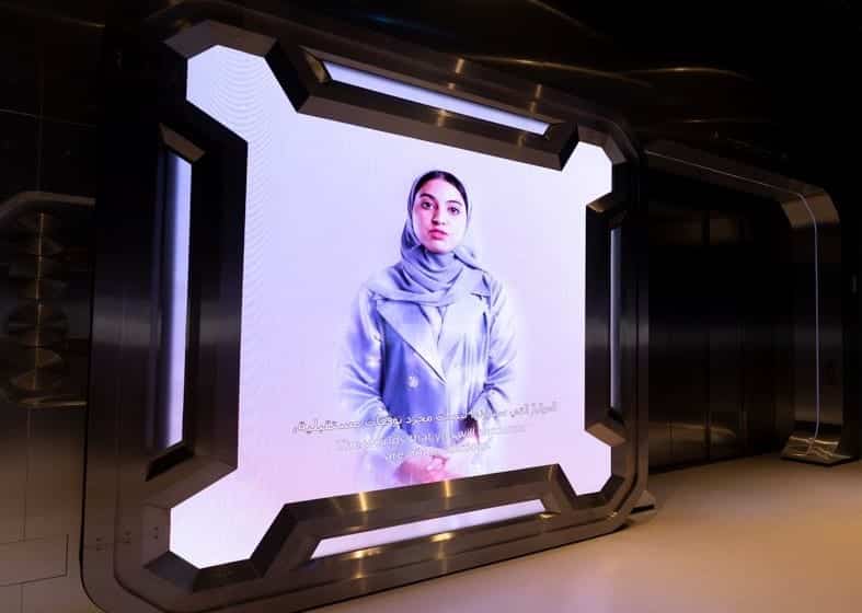 A futuristic screen shows a woman in a hijab and light coat, set in a dark space with metallic edges.