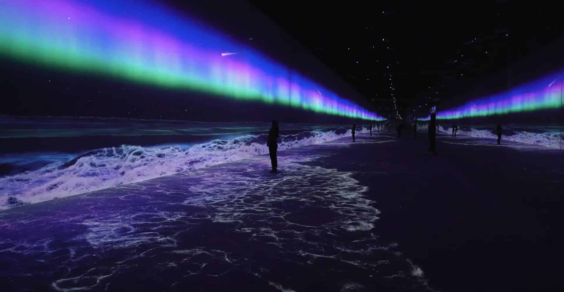 A person in a dark room with walls projecting ocean waves and auroras; the floor resembles a shoreline, creating an immersive scene.