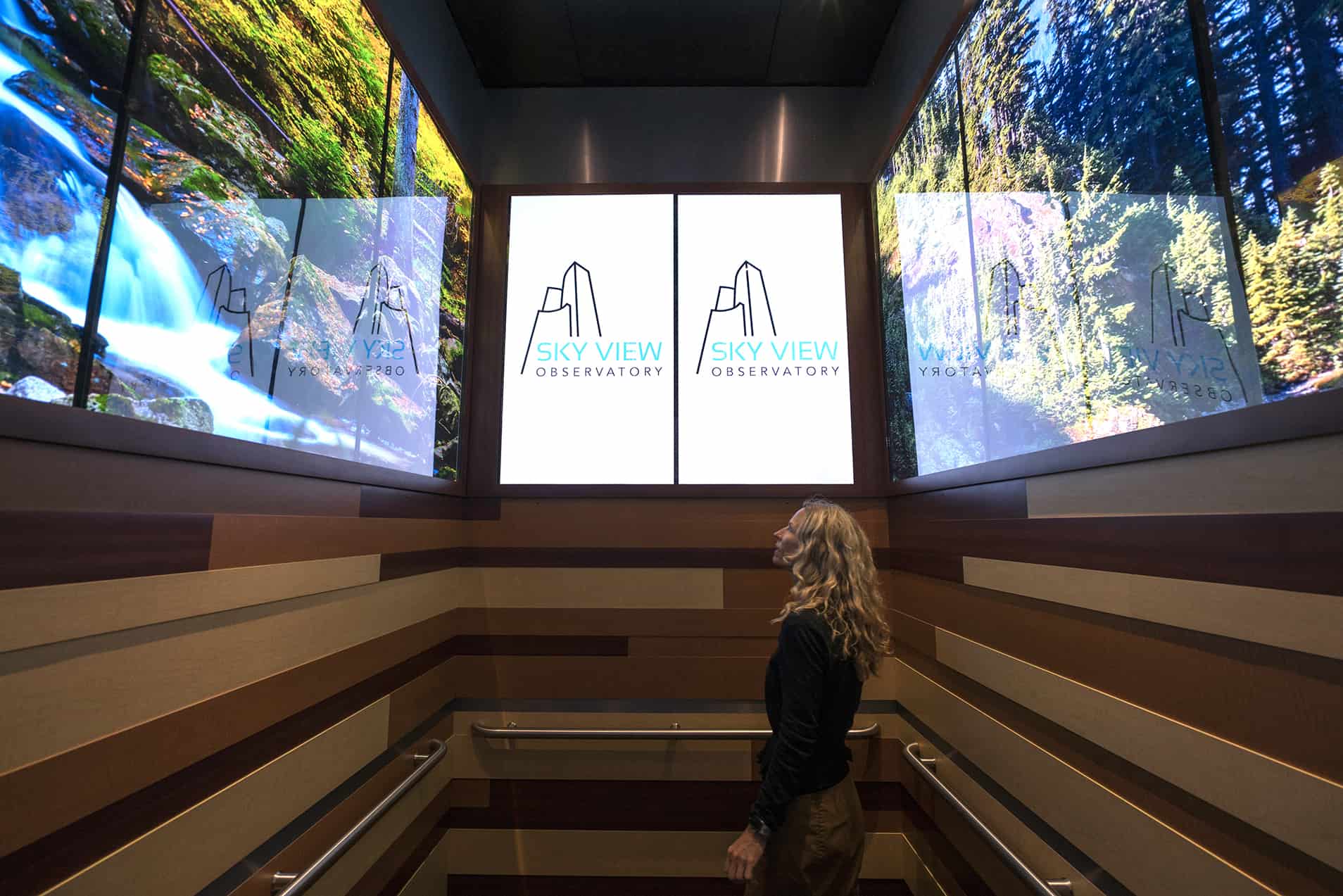 A person stands in an elevator with nature scenes on screens, Sky View Observatory logos, and brown wood paneling.