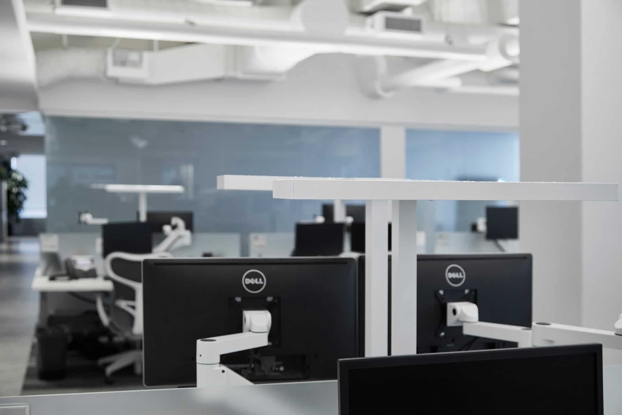 A modern office with Dell workstations, white desks, ergonomic chairs, ceiling ducts, and large windows in a minimalistic design.