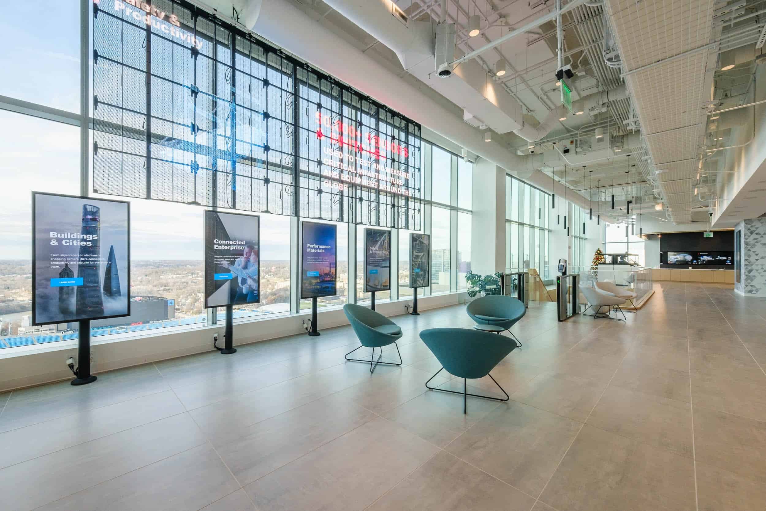 Modern office lobby with large windows, city view, circular chairs facing digital displays; exposed beams and lights; reception area behind.