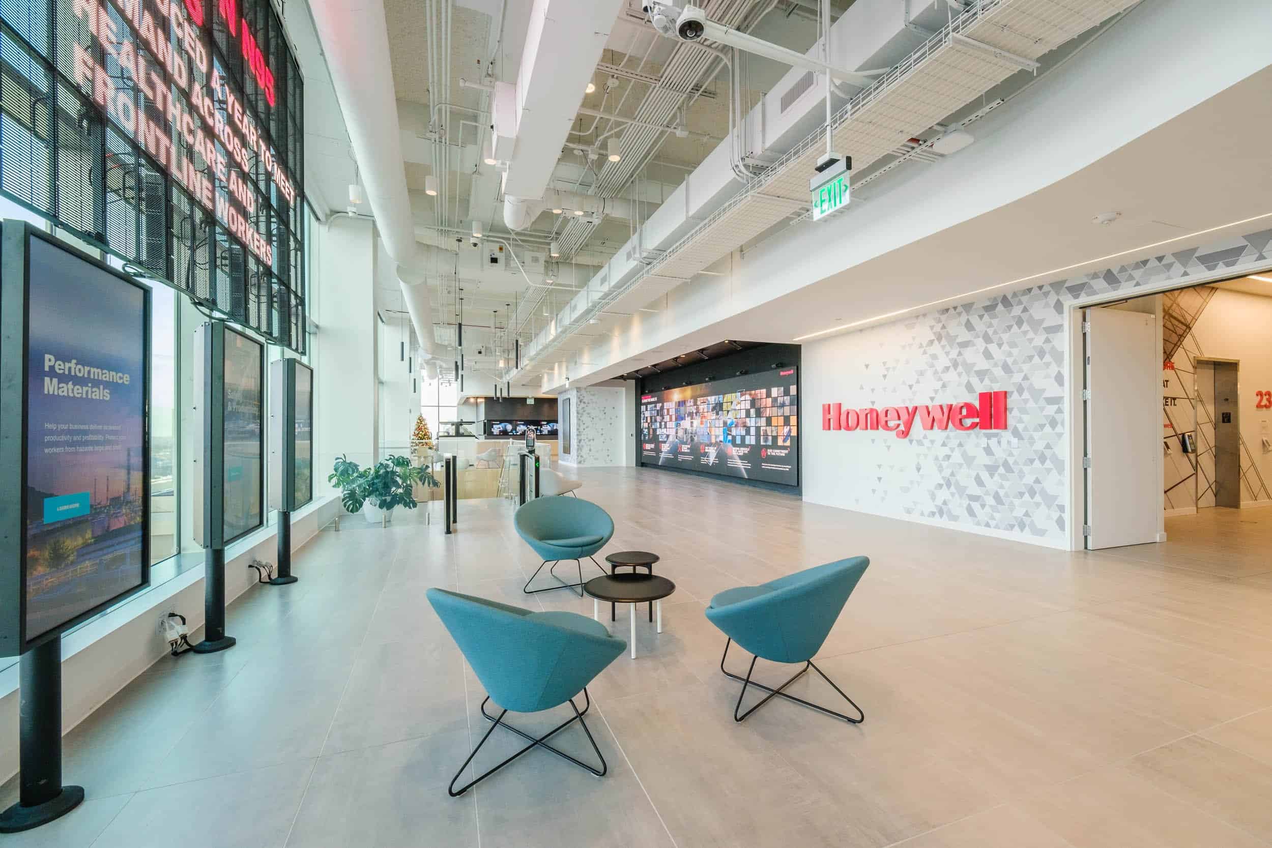 Modern lobby: light wood floors, blue chairs, black table, digital displays, Honeywell logo wall, bright lighting, open ceiling.