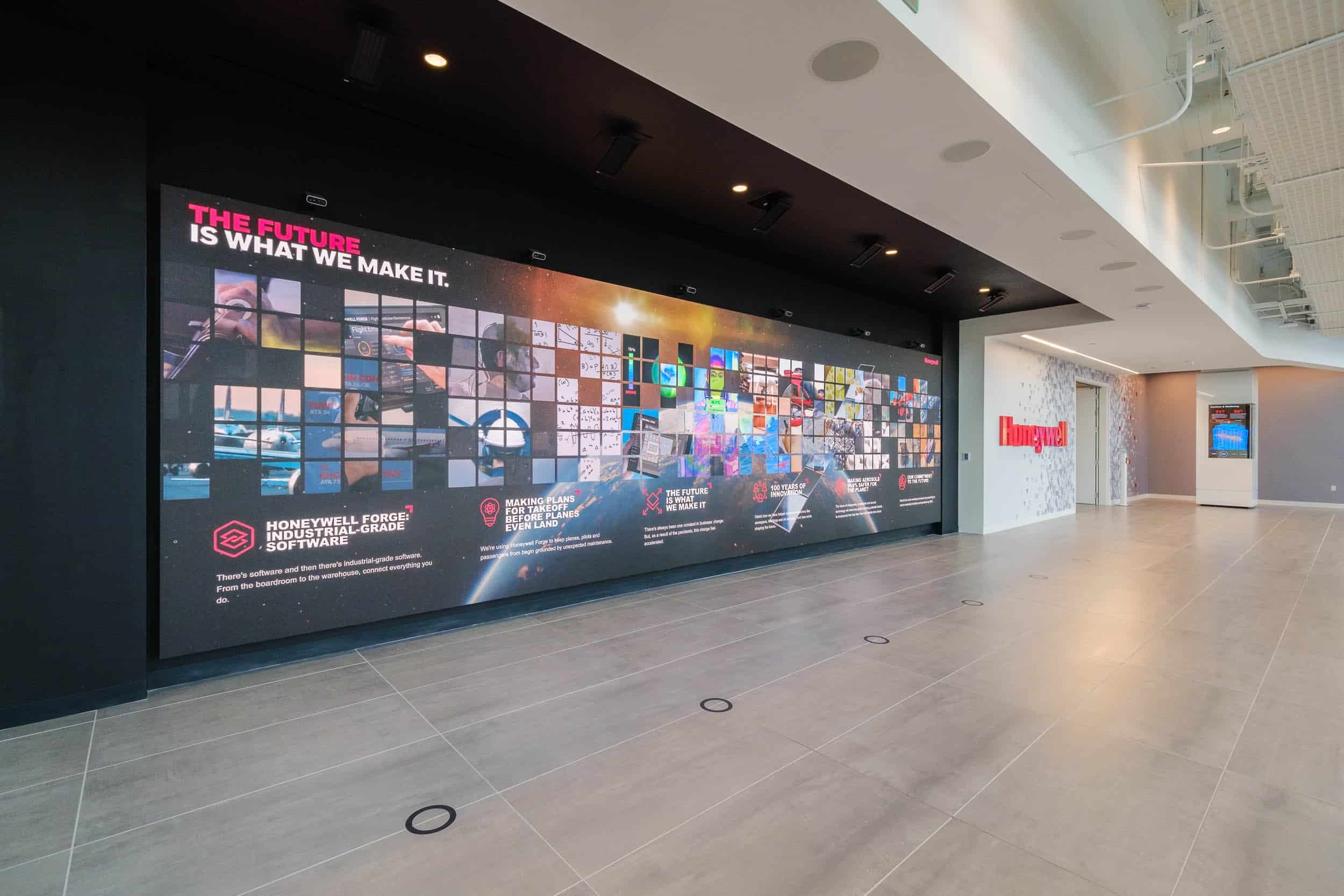 Modern office hallway with a digital wall displaying The future is what we make it. Tiled floor, company name in red on distant wall.