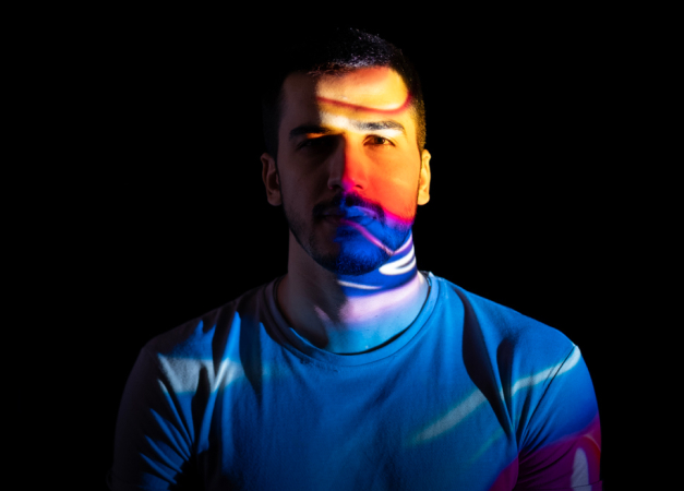 Person in a white shirt against a dark background with colorful light patterns projected onto their face and body.