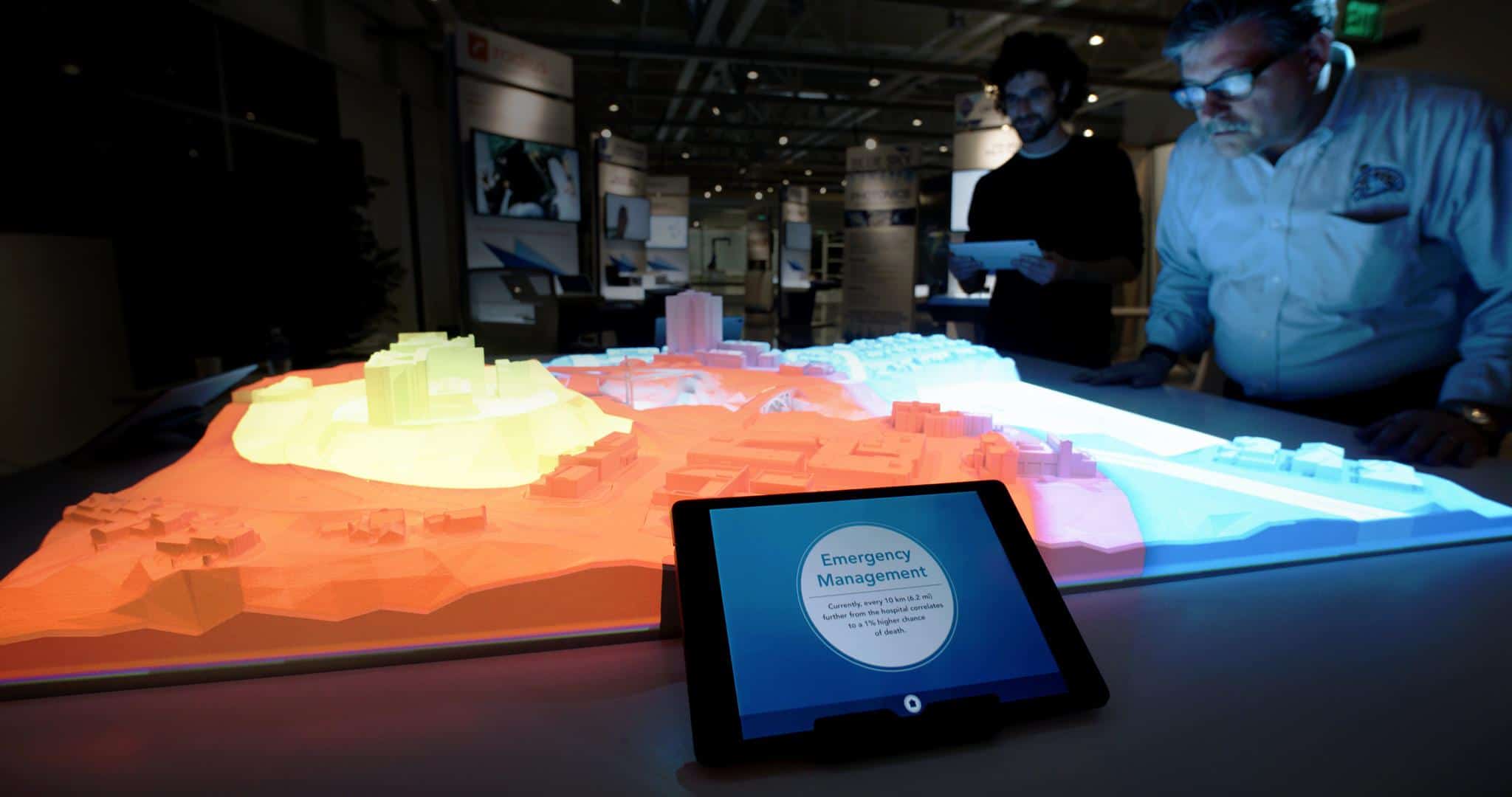 3D topo map lit with red, yellow, blue lights; two people in background examine it. Tablet in foreground reads Emergency Management.