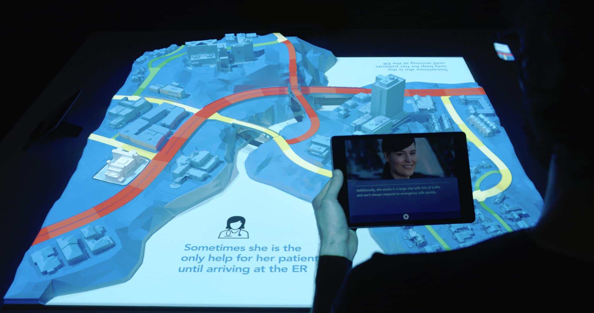 A person holds a tablet showing a smiling woman over a 3D city map with blue structures and red roads. Text reads: Shes often the only help for her patients until reaching the ER.