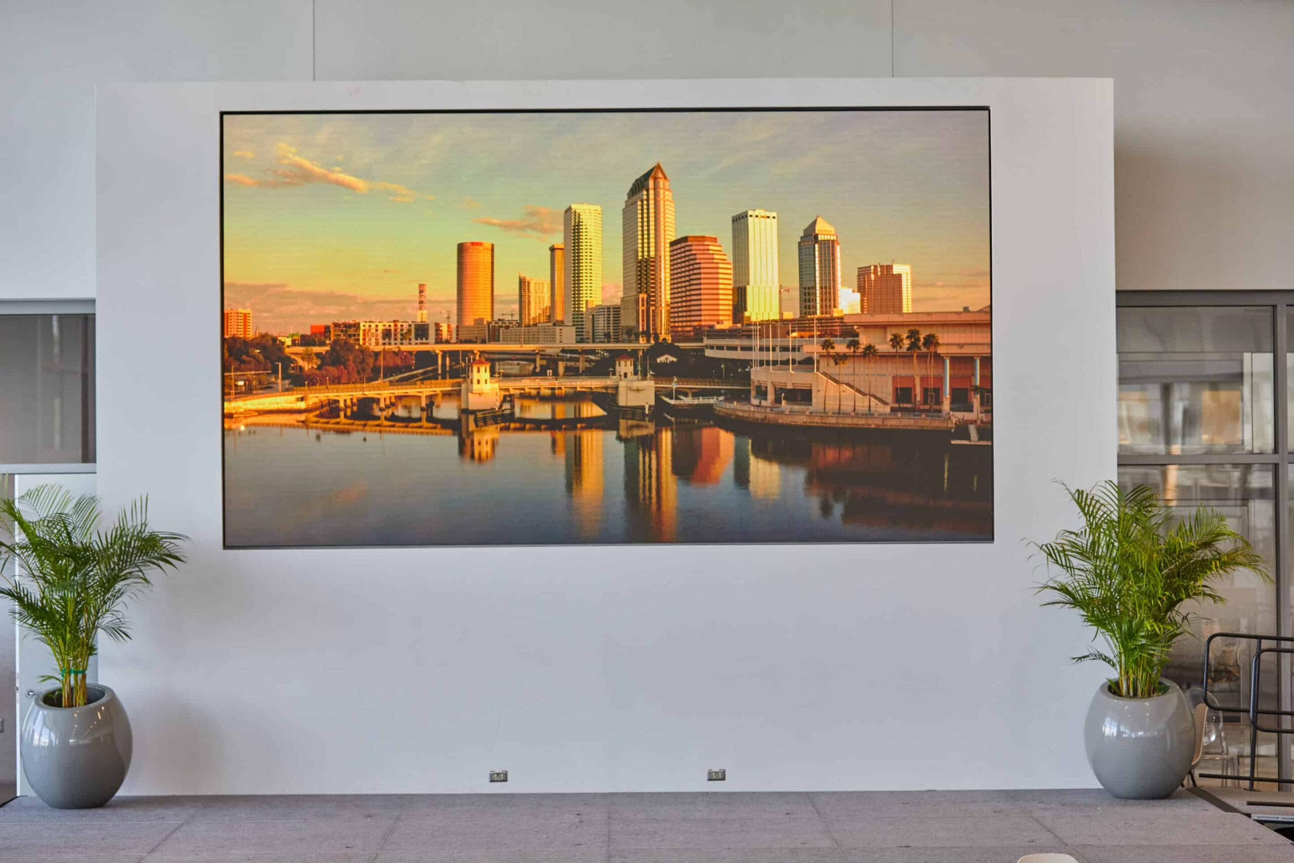 Large screen shows a sunset city skyline reflecting on water, flanked by two potted plants on a white wall.