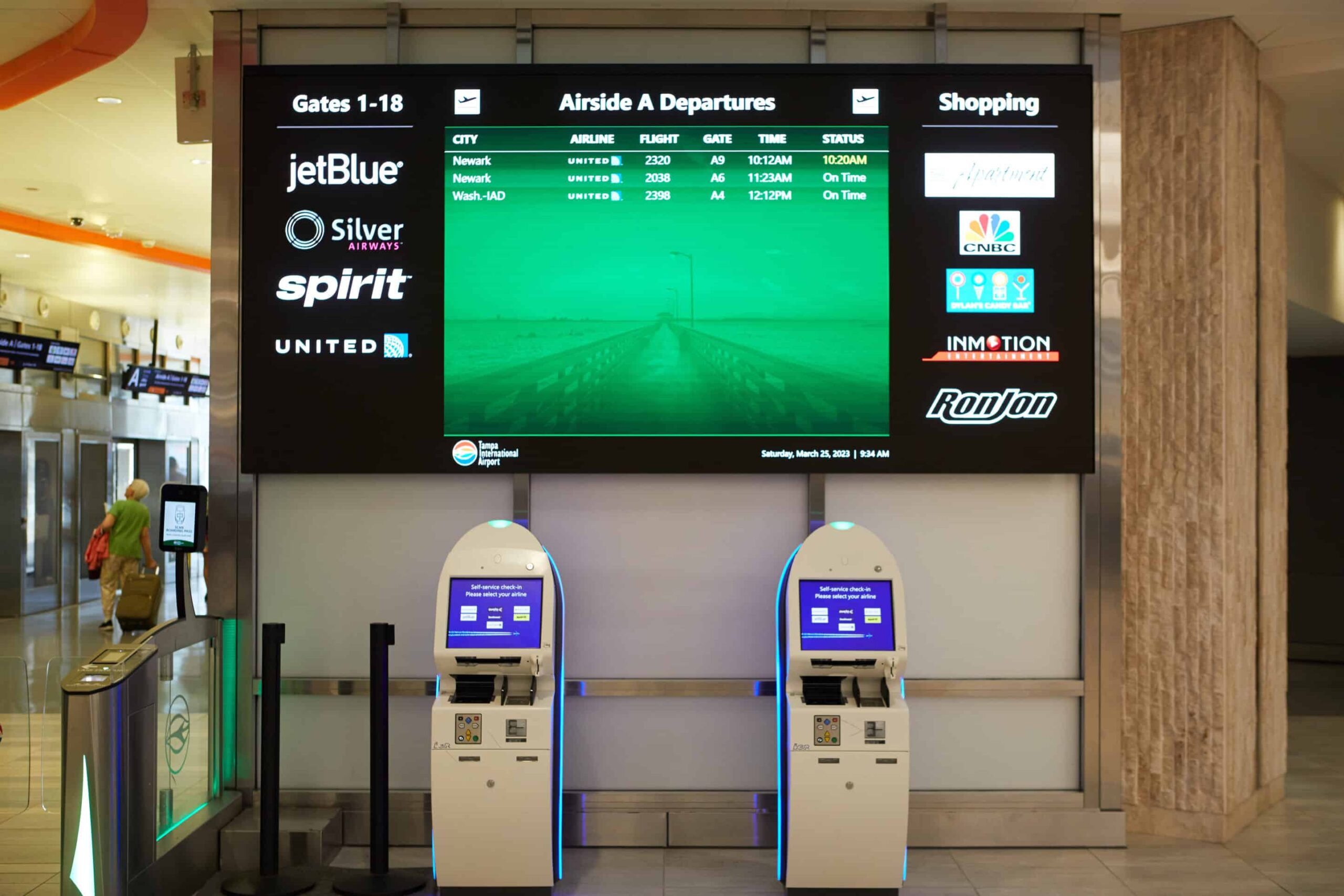 Airport info board for gates 1-18, kiosks below, bright area with airline logos and news channels on display.