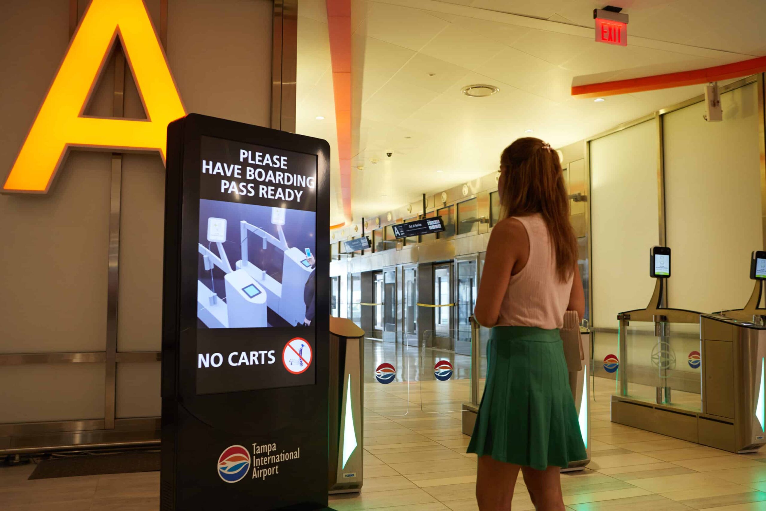 Woman at Tampa Airport digital sign: Have boarding passes ready. No carts allowed. Check-in kiosks in background.