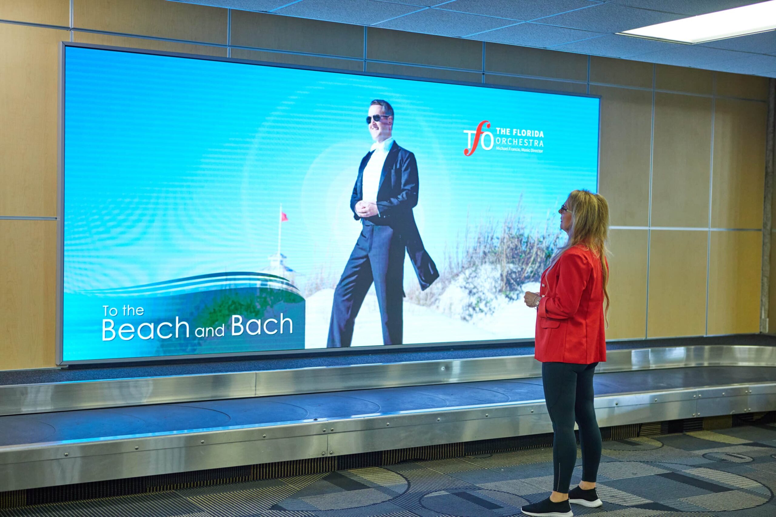 Woman in red jacket and black leggings stands by airport ad for The Florida Orchestra: To the Beach and Bach.