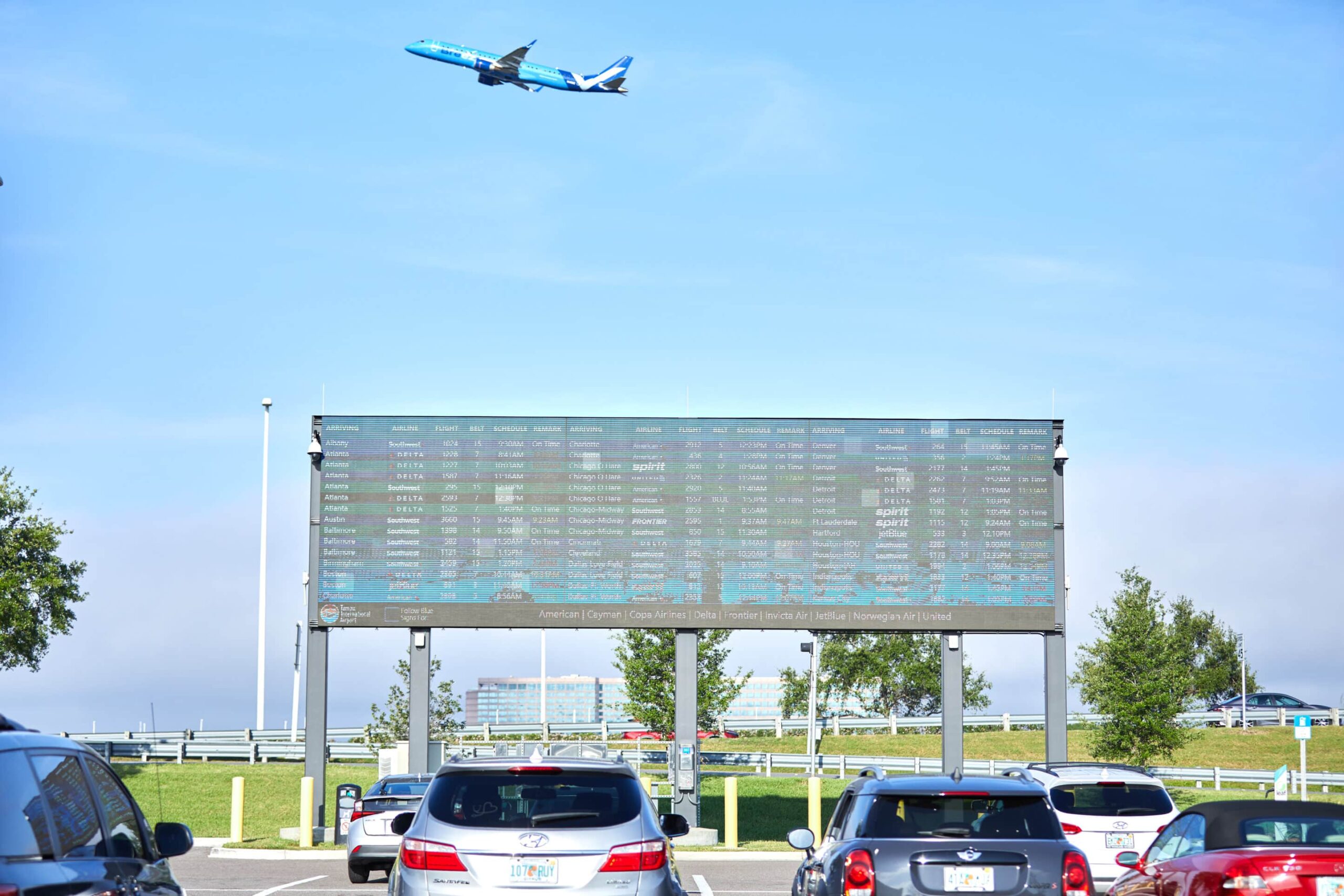 Large airport departure board outside, cars parked below; blue airplane takes off in clear sky above.