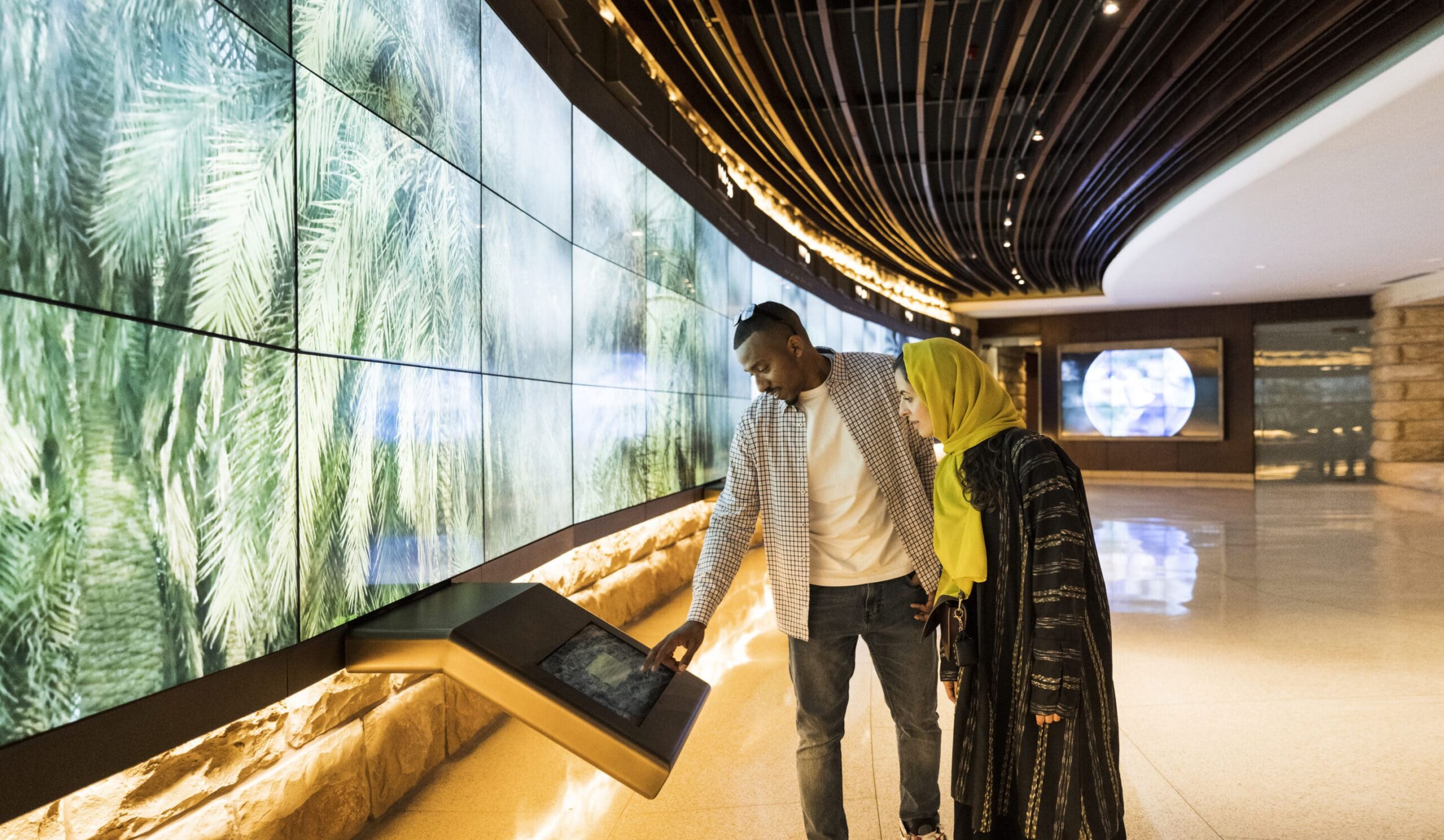 A man and woman at a modern gallery touchscreen; video of palm trees on the wall. Shes in a yellow headscarf and black dress. Sleek space.