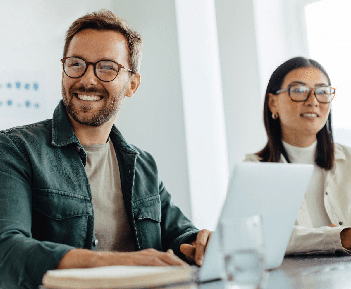 Man and woman with glasses smiling at a table; man in green jacket, woman in white blouse, laptop and glass on table.