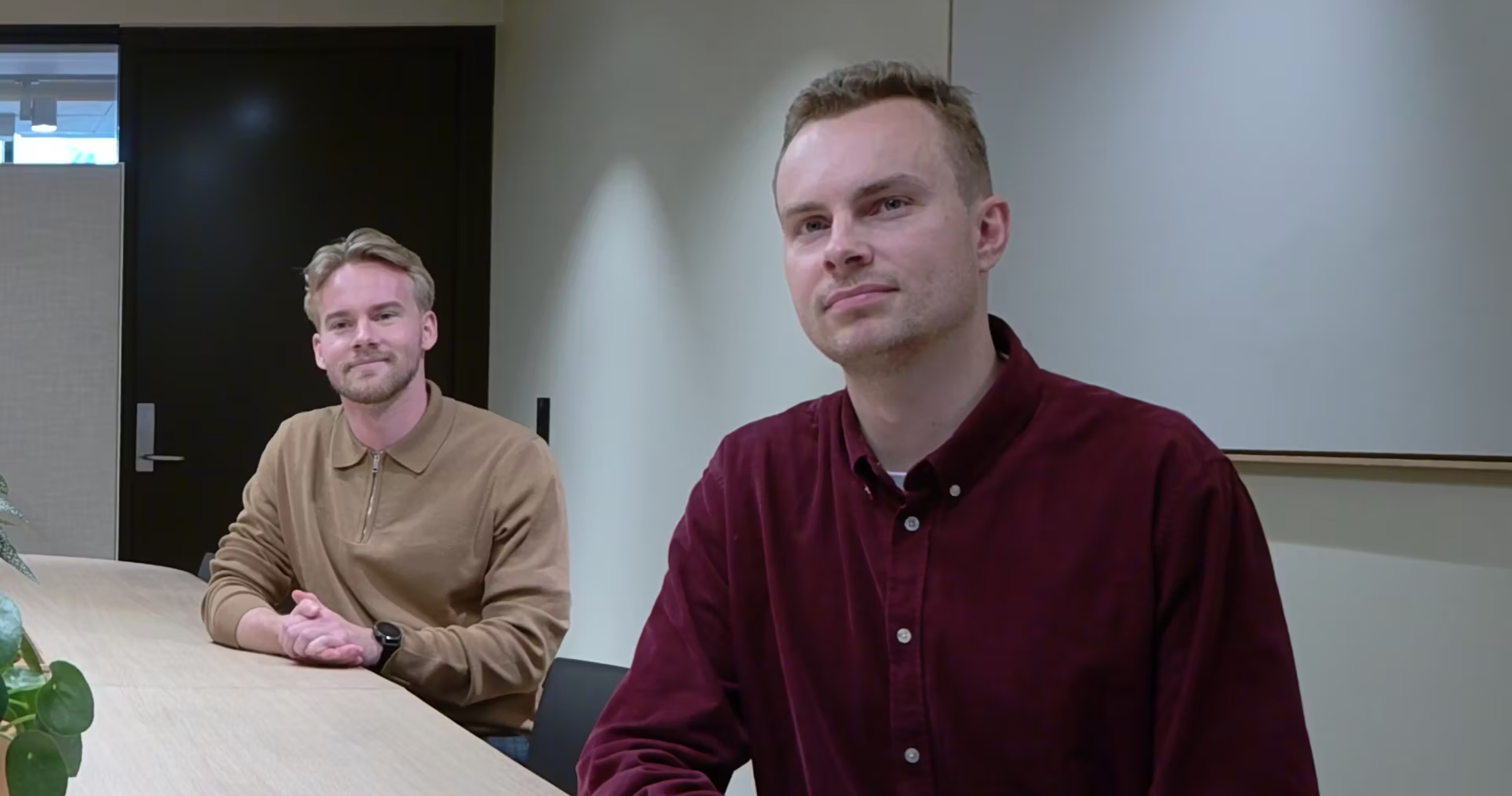 Two people at a modern office table, one in brown, the other in burgundy, facing the camera. Potted plant on the tables left.