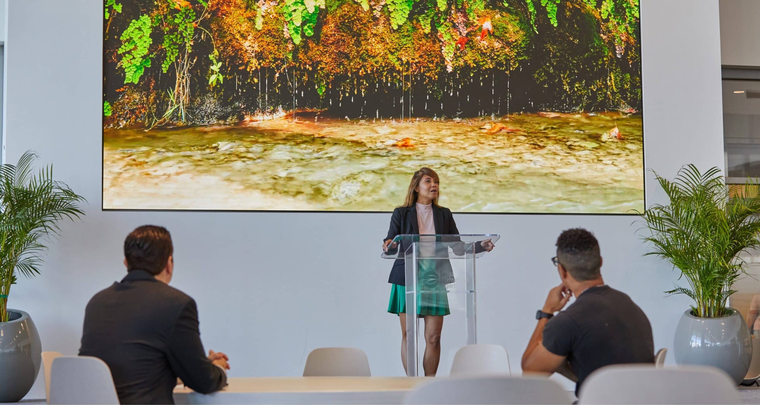 A woman in a green skirt and black blazer speaks at a podium, with nature scenery on screen; two people listen attentively.